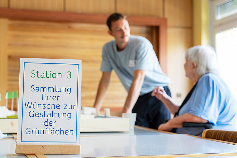 Eine ältere Frau sitzt an einem Tisch in einem Raum und lässt sich von einem Mitarbeiter von degewo beraten. Auf einem Tischaufsteller steht Station 3, Sammlung Ihrer Wünsche zur Gestaltung der Grünflächen.