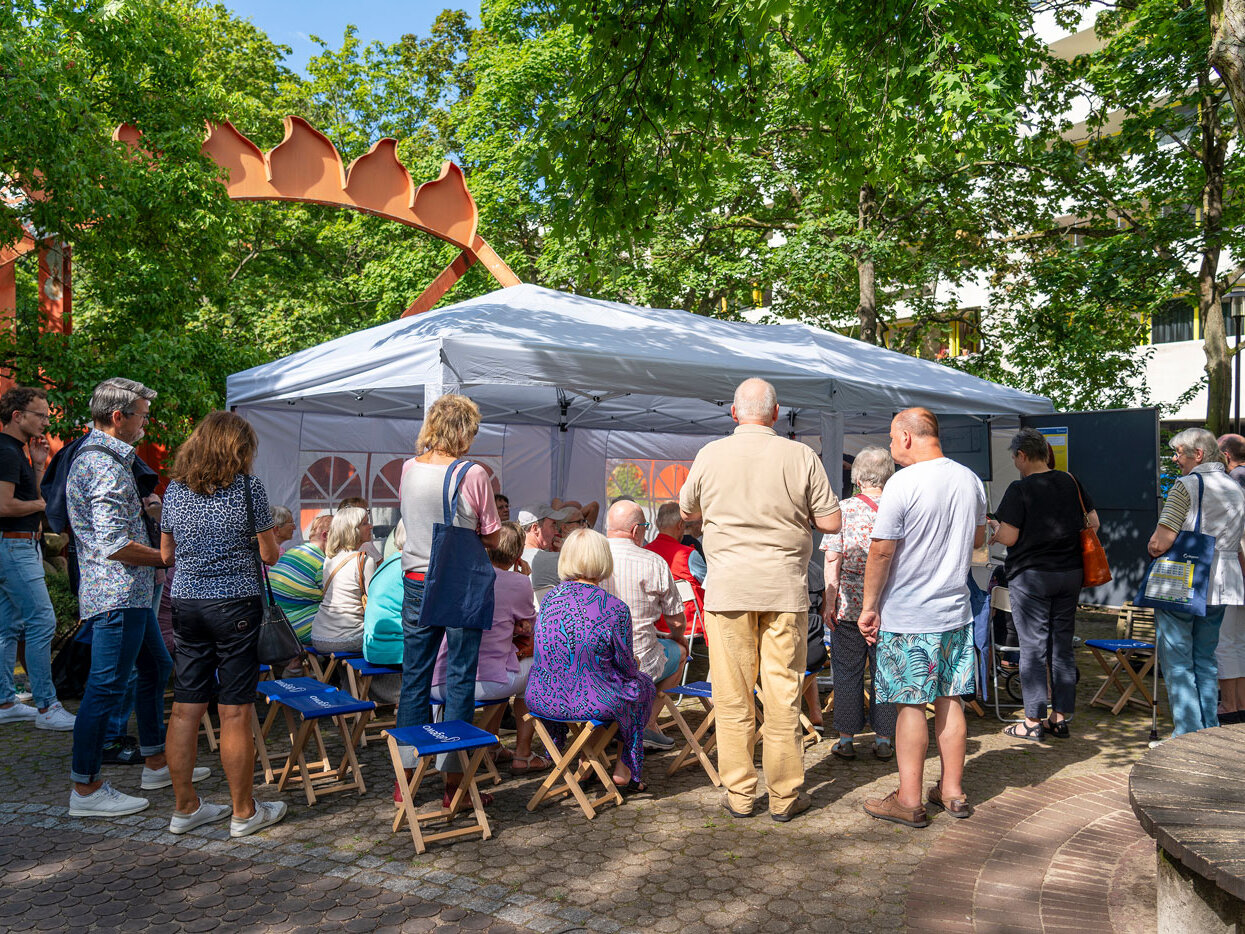 Menschen stehen und sitzen in mehreren Reihen unter einem draußen aufgestellten Pavillon. 