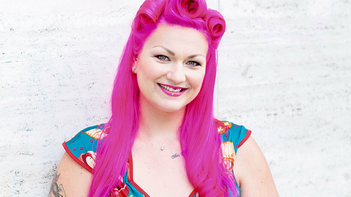 Die Konditorin Franziska Schmidt steht vor einer hellgrauen Wand und lächelt in die Kamera. Ihre Haare sind auffallend pink, ihr blaues Kleid ist mit roten Cupcakes und einer Schleife verziert.