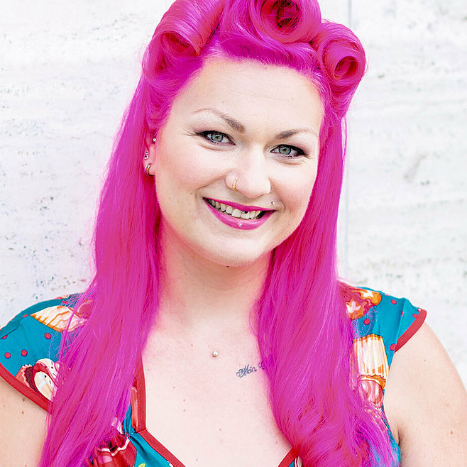 Die Konditorin Franziska Schmidt steht vor einer hellgrauen Wand und lächelt in die Kamera. Ihre Haare sind auffallend pink, ihr blaues Kleid ist mit roten Cupcakes und einer Schleife verziert.