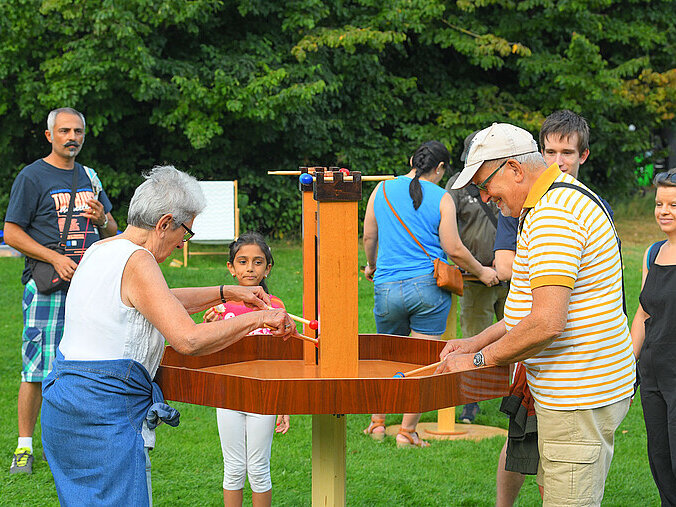 Zwei ältere Menschen beschäftigen sich mit einem XXL-Holzspiel, das auf dem Rasen steht. Andere Gäste schauen ihnen dabei zu.