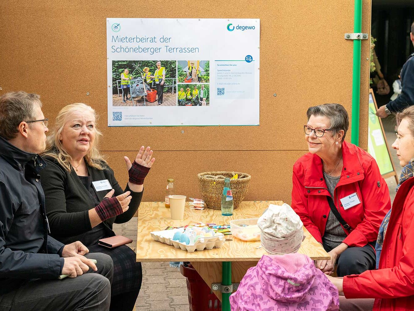 Gesprächsrunde an einem Infotisch bei der degewo-Informationsveranstaltung in den Schöneberger Terrassen. Vier Erwachsene und ein Kind sitzen zusammen, diskutieren und informieren sich. Im Hintergrund hängt ein Plakat zum Mieterbeirat der Schöneberger Terrassen.