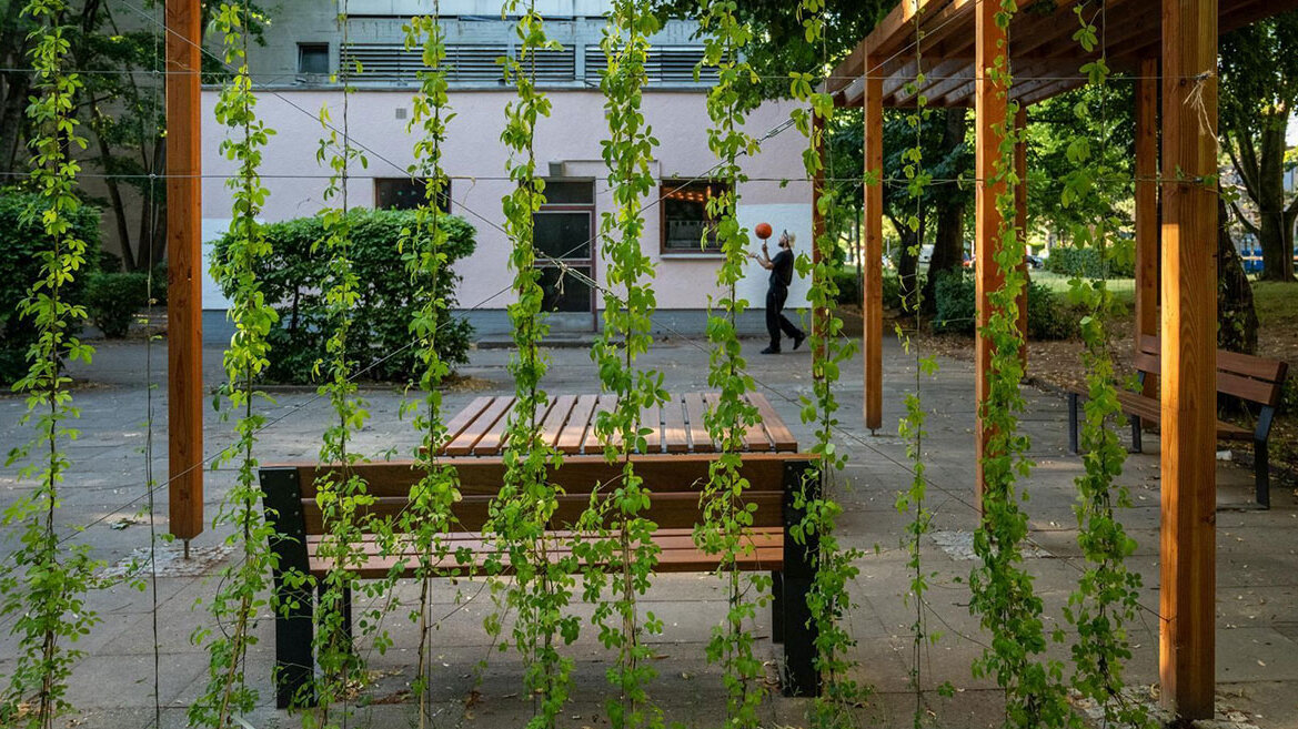 Pergola im Innenhof eines Gebäudes. Ein Mann spielt Basketball im Hintergrund.