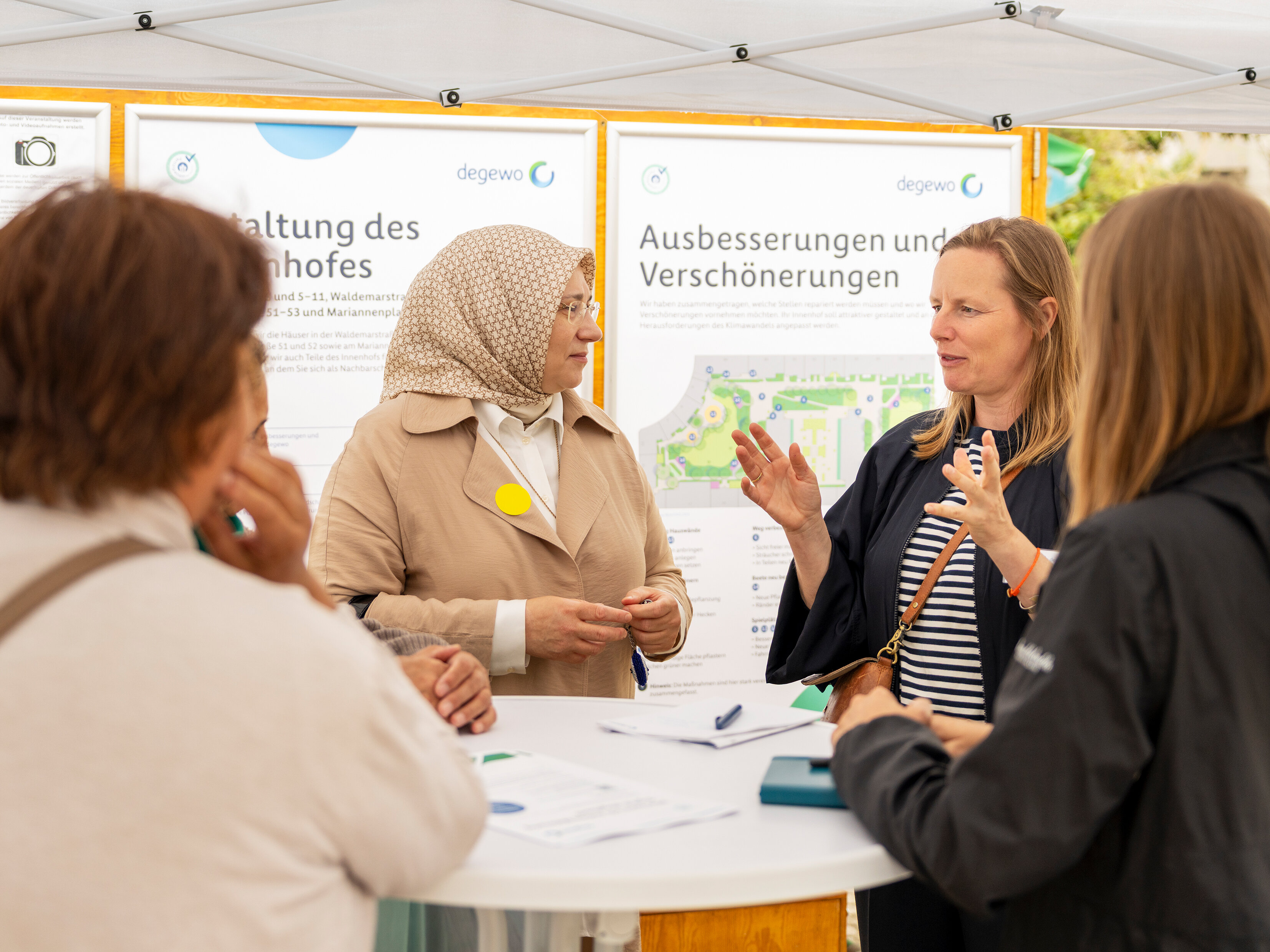 Mehrere Personen stehen um einen runden Tisch und unterhalten sich. Im Hintergrund zwei große Plakate.