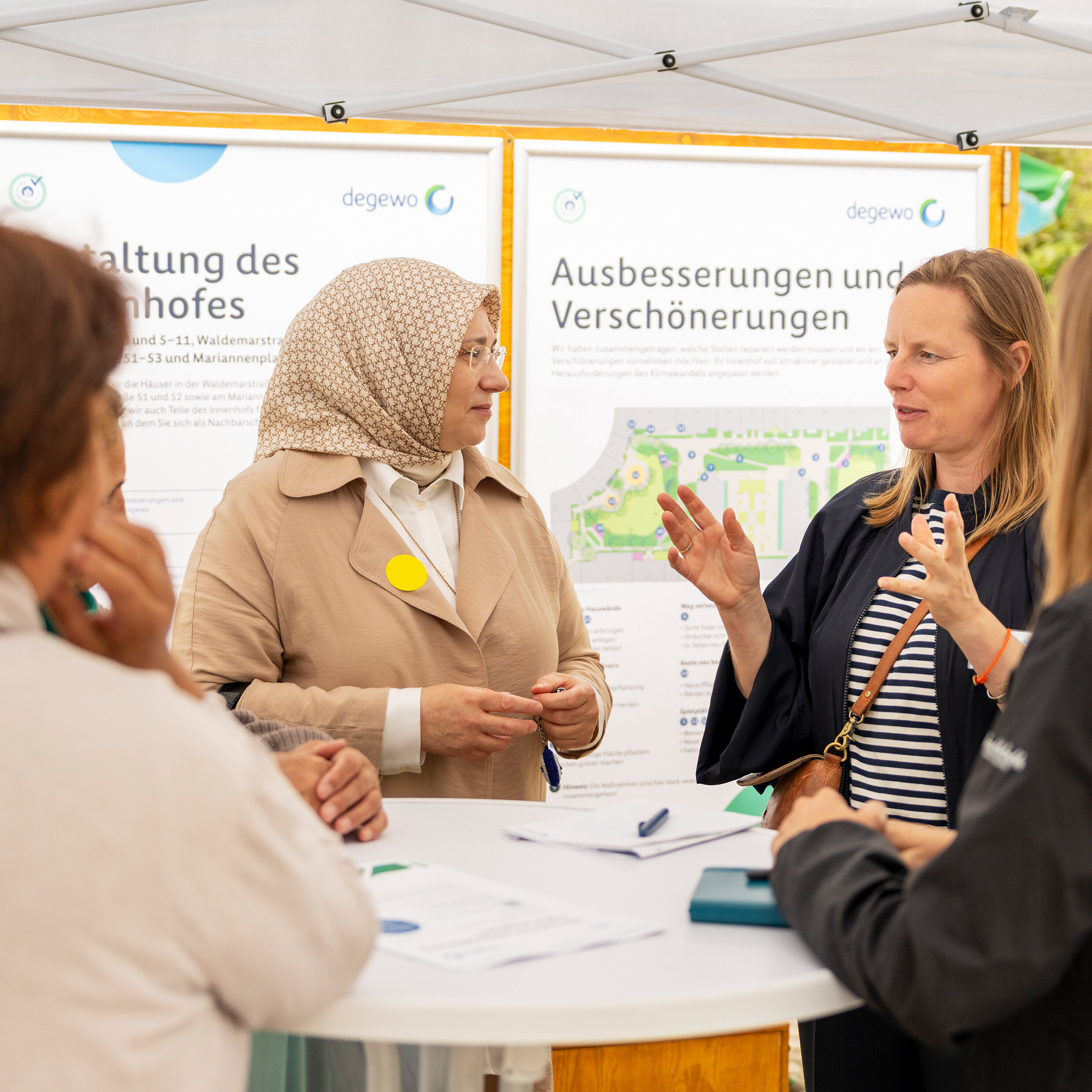 Mehrere Personen stehen um einen runden Tisch und unterhalten sich. Im Hintergrund zwei große Plakate.