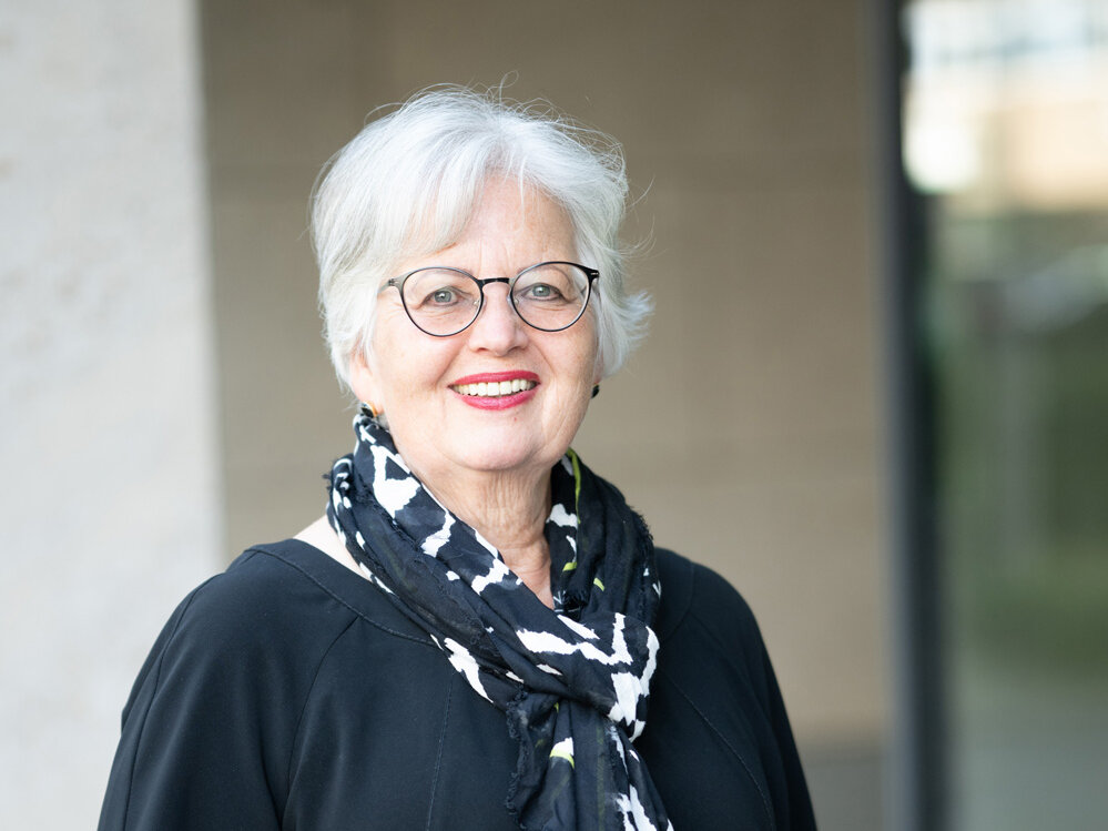 Ältere Frau mit weißem Haar und Brille trägt ein schwarzes Kleid und einen gemusterten Schal und steht im Freien vor einem Gebäude; sie lächelt und blickt in die Kamera.