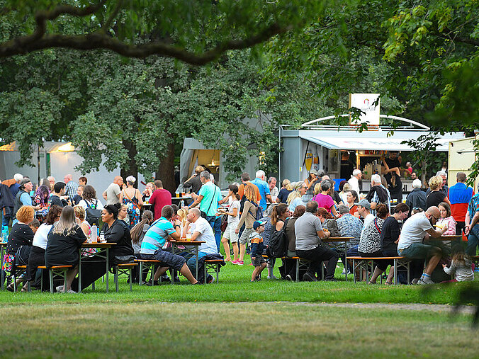 Zu sehen sind Gäste, die in einem Park an Tischen sitzen und essen und trinken. Im Hintergrund sind Verkaufsstände zu sehen. 