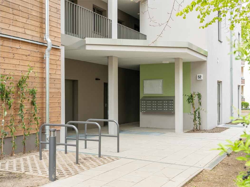 Außenansicht degewo-Neubau Beyrodtstraße 52: Eingangsbereich mit Weg, Weglaterne und Fahrradständern sowie rechts Baumblättern im Vordergrund.