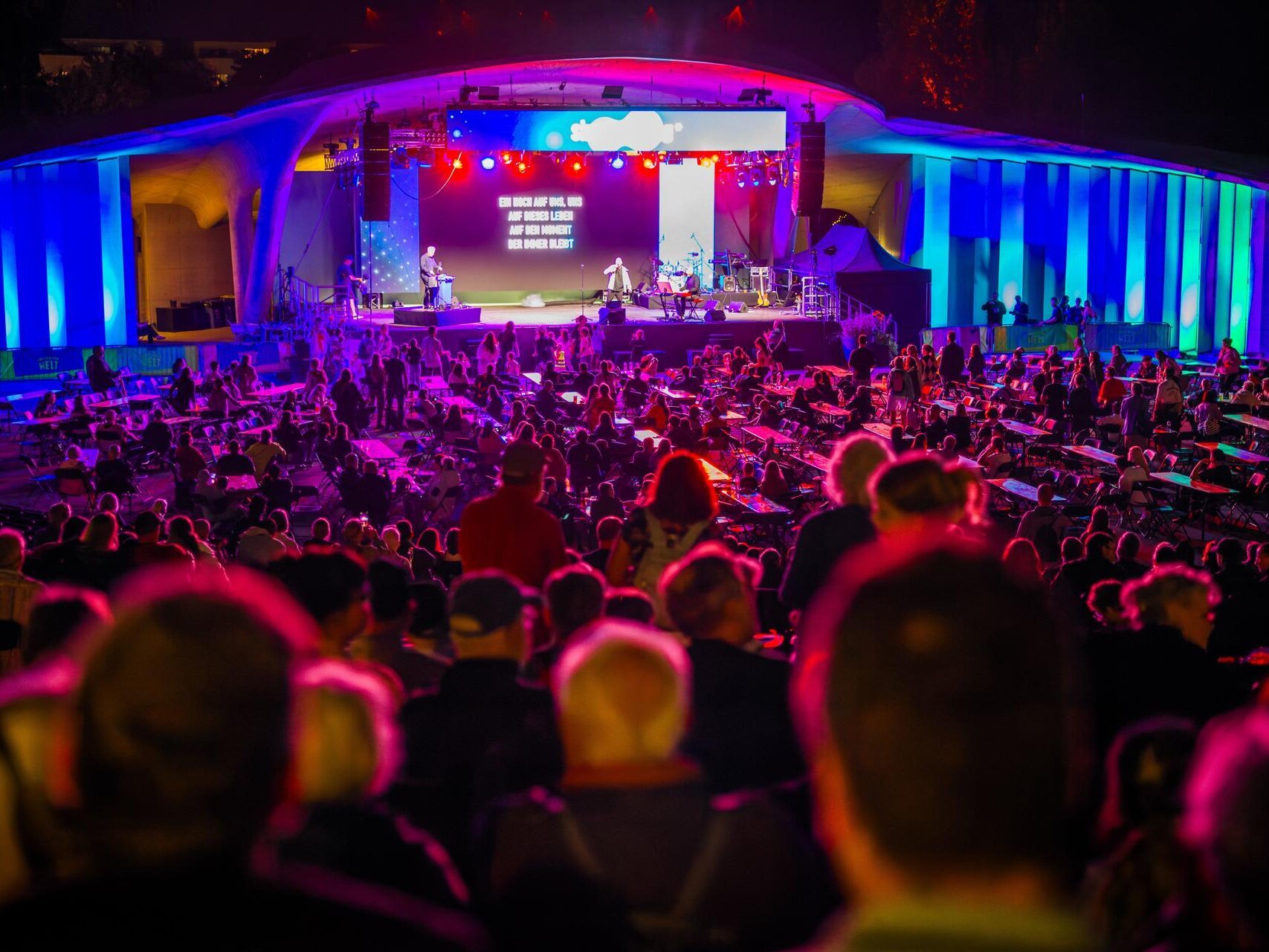 Blick über ein voll besetztes Freiluft-Amphitheater auf eine farbenfroh beleuchtete Bühne. Zahlreiche Besucherinnen und Besucher verfolgen das Abendprogramm von den Sitzplätzen und Tischen aus.