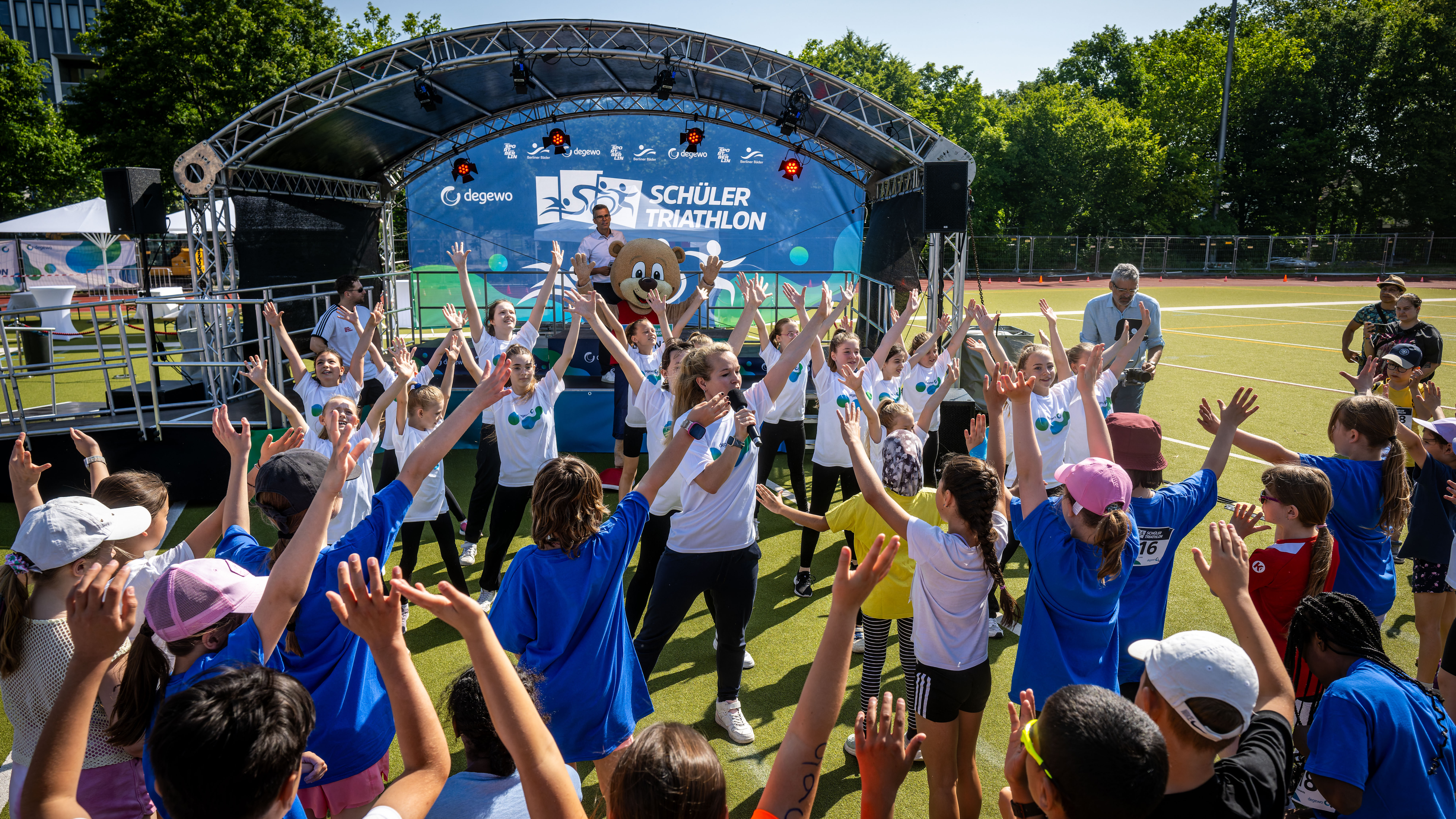 Viele Kinder stehen vor einer Bühne im Freien und heben die Arme beim Mitmachen.