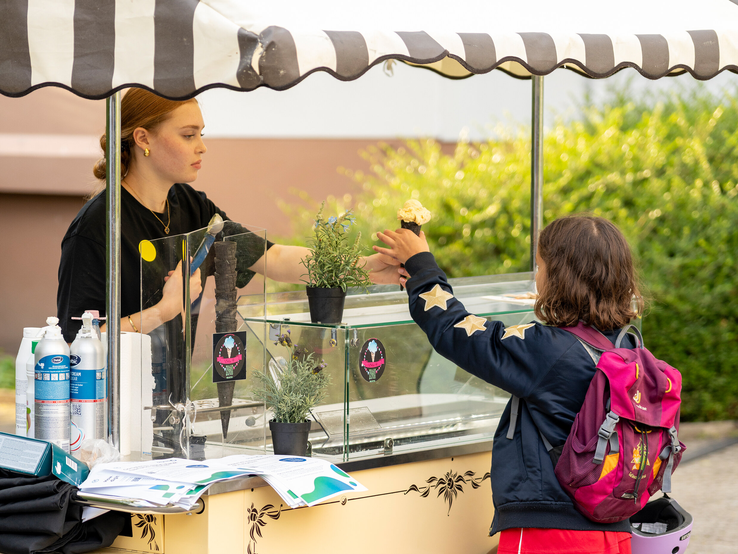 Kind kauft an einem mobilen Eisstand ein; Verkäuferin reicht eine Eiswaffel über die Theke, daneben Pflanzen und Informationsmaterial.