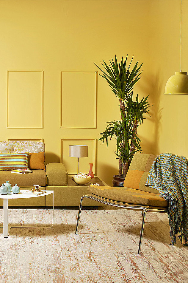 Ein Wohnzimmer mit senfgelber Couch, Couchtisch, Sessel und diversen Accessoires. Im Fokus steht die gelbe Wandfarbe, die sich über die gesamte Wandfläche verteilt.