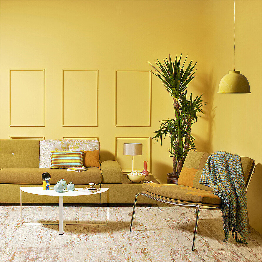 Ein Wohnzimmer mit senfgelber Couch, Couchtisch, Sessel und diversen Accessoires. Im Fokus steht die gelbe Wandfarbe, die sich über die gesamte Wandfläche verteilt.