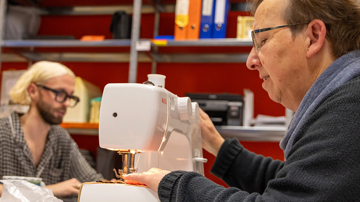 Eine Mitarbeiterin sitzt an der Nähmaschine, im Hintergrund arbeitet ein Mann am Computer.