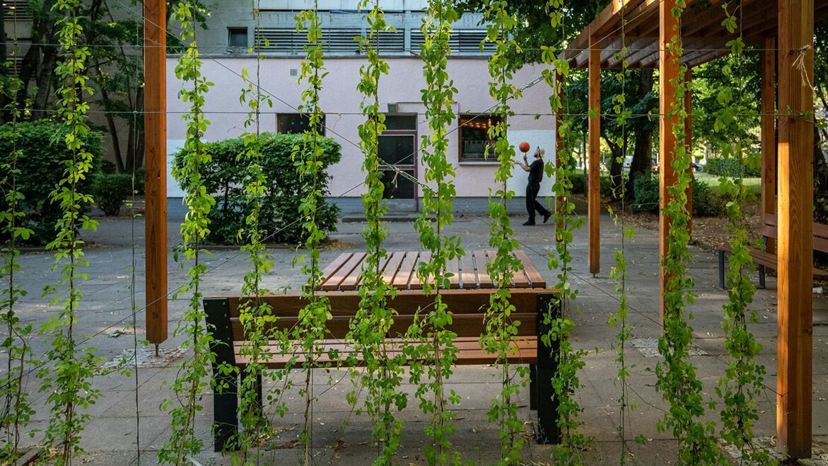 Pergola im Innenhof eines Gebäudes. Ein Mann spielt Basketball im Hintergrund.