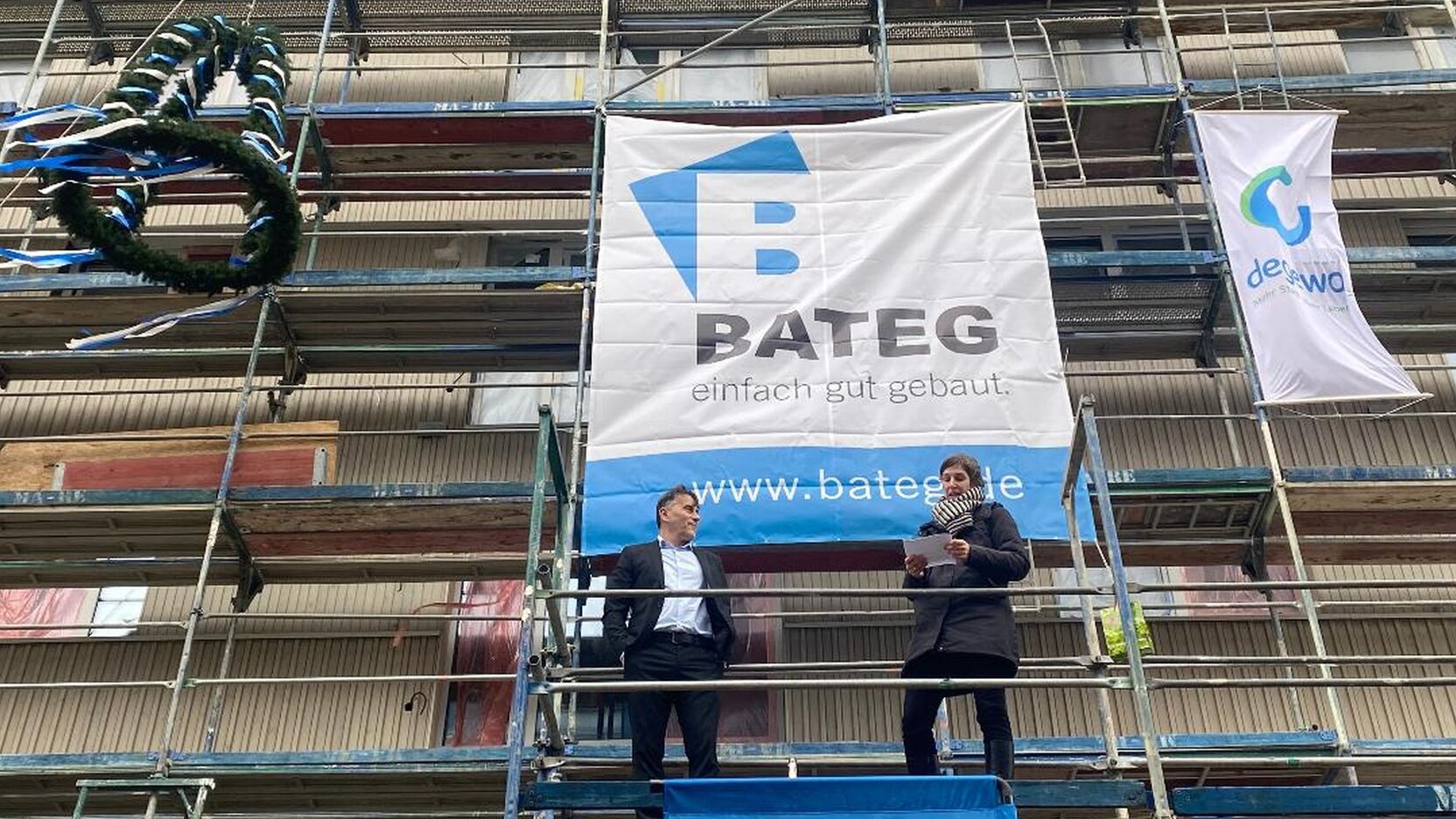 Ein Mann und eine Frau stehen auf einem Baugerüst. Oben links am Gerüst hängt eine Richtkrone.