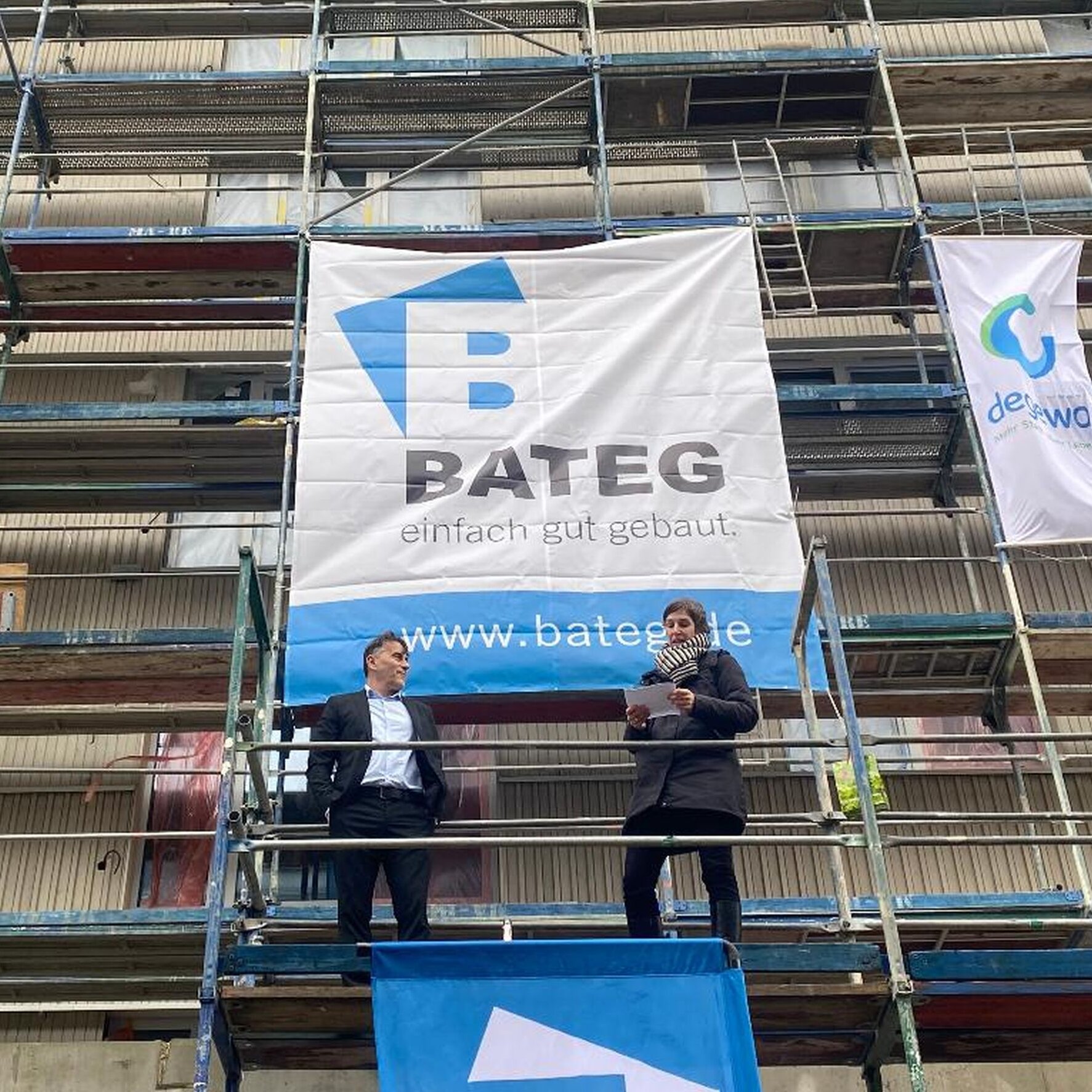 Ein Mann und eine Frau stehen auf einem Baugerüst. Oben links am Gerüst hängt eine Richtkrone.