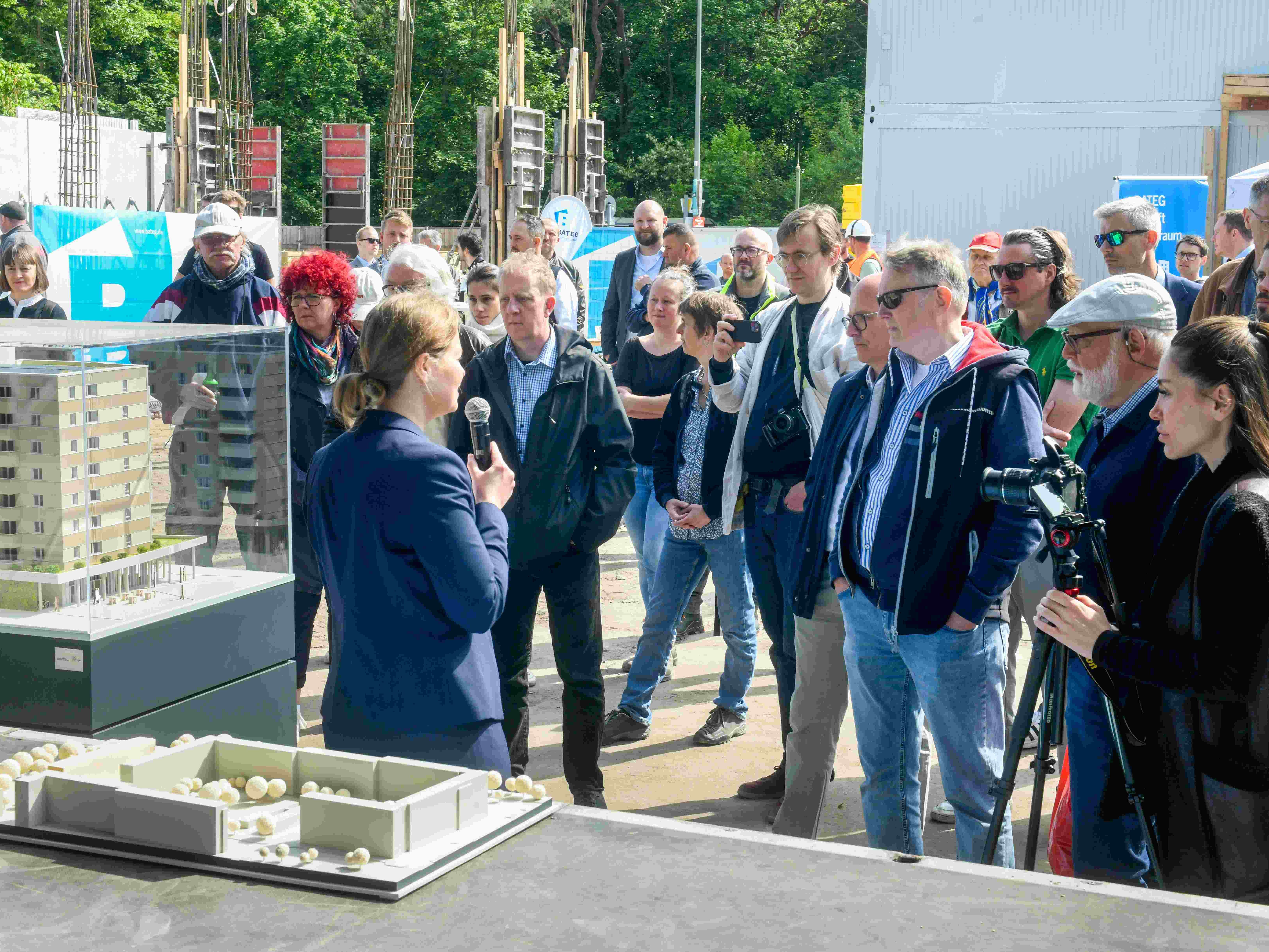 Eine Frau mit Mikrofon spricht vor einer Gruppe von etwa 30 Personen auf einer Baustelle. Vor ihr stehen Architekturmodelle eines mehrgeschossigen Wohngebäudes in Glasvitrinen. Mehrere Zuhörende tragen Jacken, einige halten Kameras oder Smartphones. Im Hintergrund sind Baukräne, Schalungselemente und Bäume sichtbar.