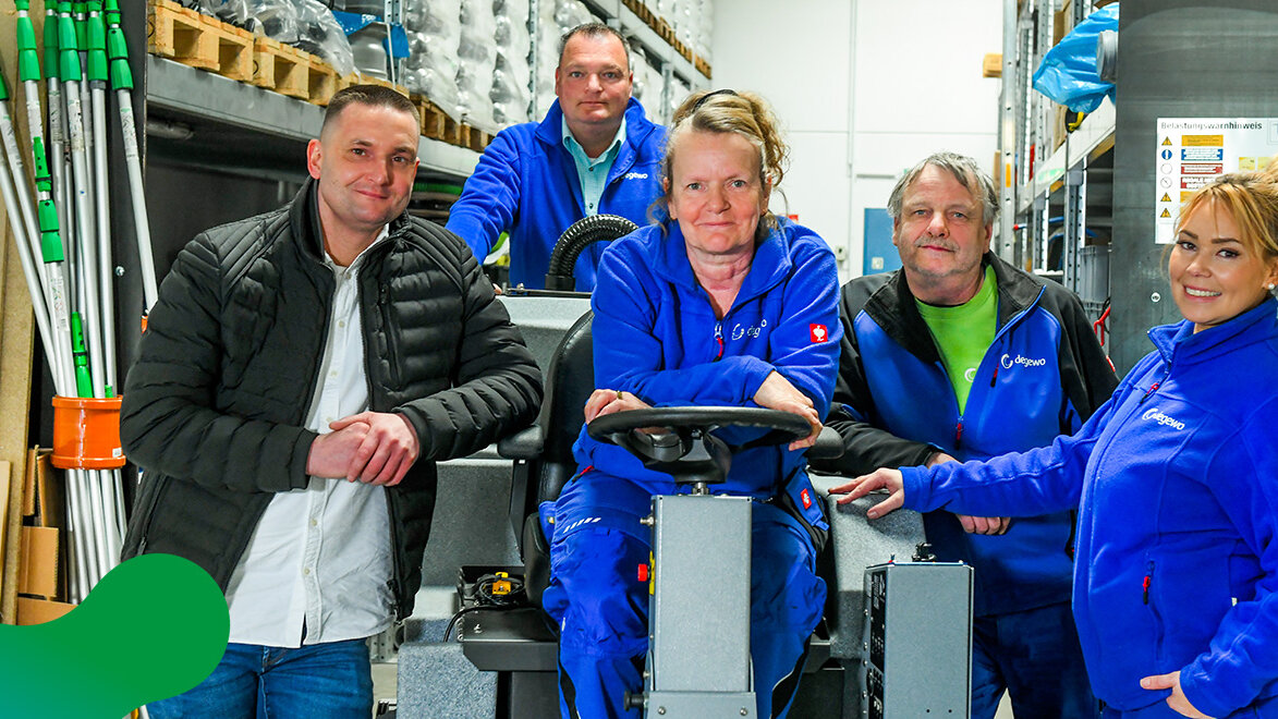 Ein Team aus fünf Mitarbeitenden posiert gemeinsam in einem Reinigungs- und Lagerbereich wobei eine Mitarbeiterin auf einer Reinigungsmaschine sitzt und alle in die Kamera lächeln.