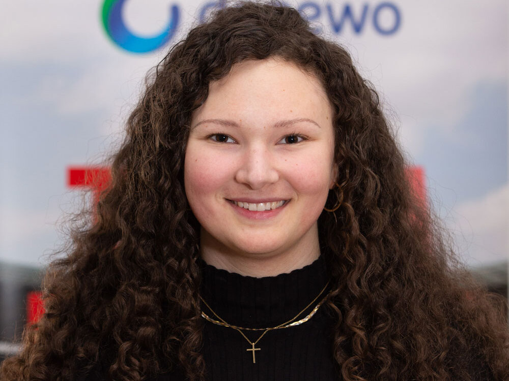 Junge Frau mit langen Locken steht vor einer Sponsorenwand mit den Logos von LOTTO Berlin und degewo und lächelt in die Kamera.