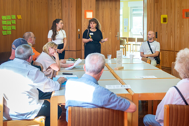 Die Anwohner eines zu sanierenden Wohngebäudes sitzen an einem Tisch und hören den Mitarbeiterinnen zu. Diese stehen am Ende des Tisches und informieren.