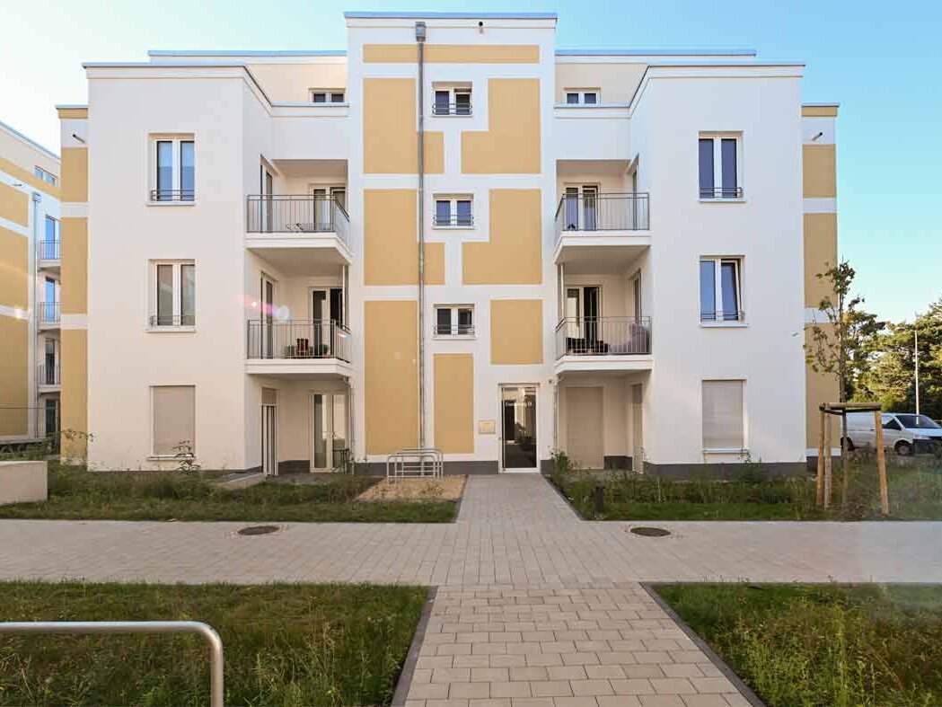 Frontalansicht eines degewo-Neubaus am Eisenhutweg in Berlin. Das dreigeschossige Mehrfamilienhaus mit heller Fassade und gelben Akzentflächen verfügt über Balkone und einen zentralen Eingangsbereich. Ein gepflasterter Weg führt durch begrünte Außenanlagen zum Gebäude und unterstreicht den modernen, wohnlichen Charakter des Neubauprojekts.