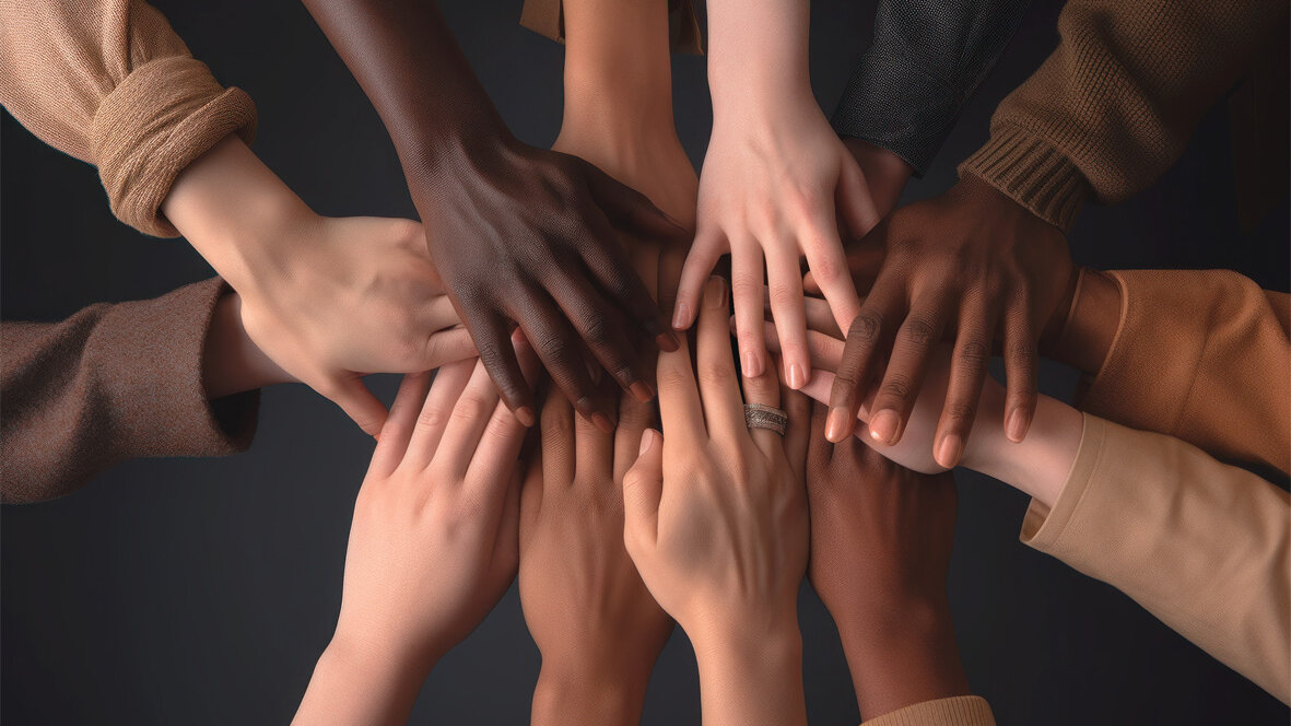 Mehrere Hände unterschiedlicher Hauttöne liegen übereinander in der Mitte des Bildes; die Personen tragen langärmelige Kleidung in neutralen Farben, der Hintergrund ist dunkel und einfarbig.