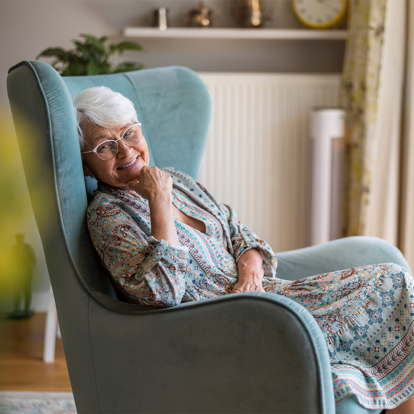Eine ältere Frau sitzt auf einem Stuhl vor dem Fenster und lächelt.