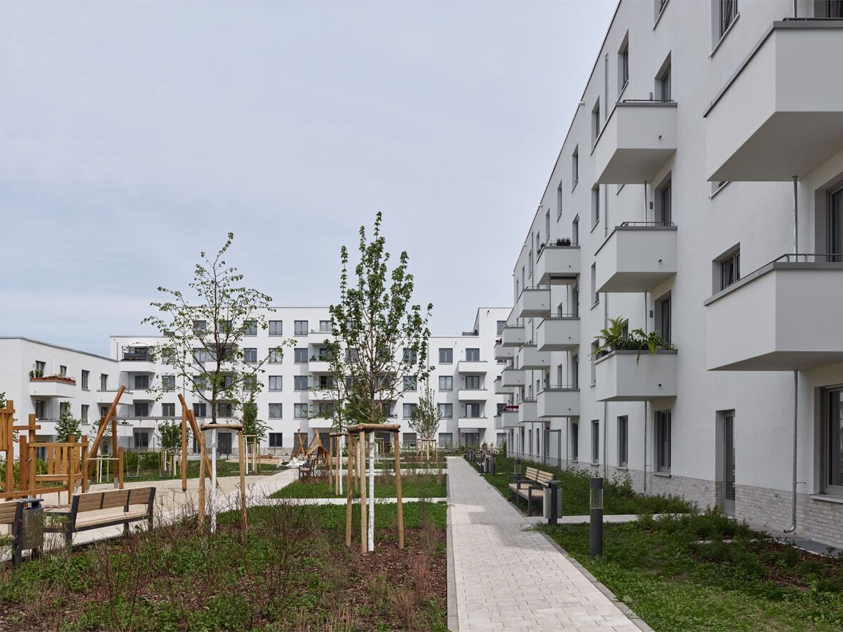 Außenansicht degewo-Neubau Dessauerstraße 39 bis 45 a-b Retzowstraße 54 bis 60: Hofseite mit Sitzbänken, Beeten, neu gepflanzten Bäumen, Weg und Weglaternen im Vordergrund.