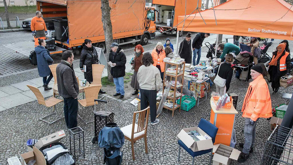 Menschen stöbern an Ständen unter einem orangefarbenen BSR-Pavillon. Rundherum stehen Möbel, Bücher und Haushaltsgegenstände zum Mitnehmen und Weitergeben bereit.