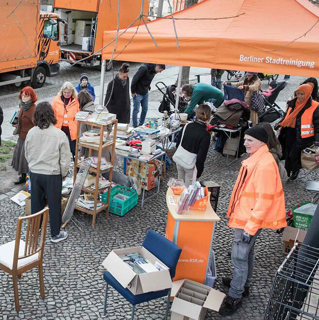 Menschen stöbern an Ständen unter einem orangefarbenen BSR-Pavillon. Rundherum stehen Möbel, Bücher und Haushaltsgegenstände zum Mitnehmen und Weitergeben bereit.