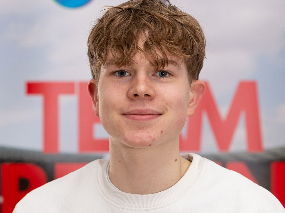 Ein junger Mann in einem weißen Sweatshirt steht vor einem LOTTO-Berlin- und degewo-Hintergrund und blickt ruhig in die Kamera.