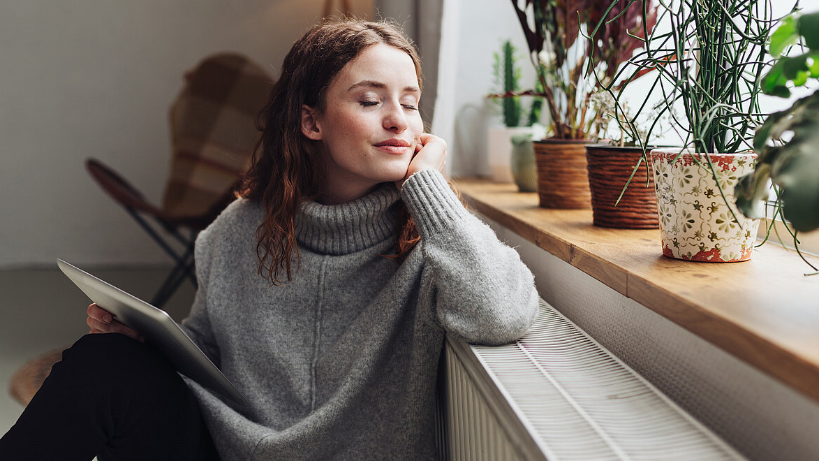 Eine Frau sitzt mit geschlossenen Augen auf dem Boden an die Heizung am Fenster gelehnt mit einem Tablet in der Hand und genießt die Sonne auf ihrem Gesicht.
