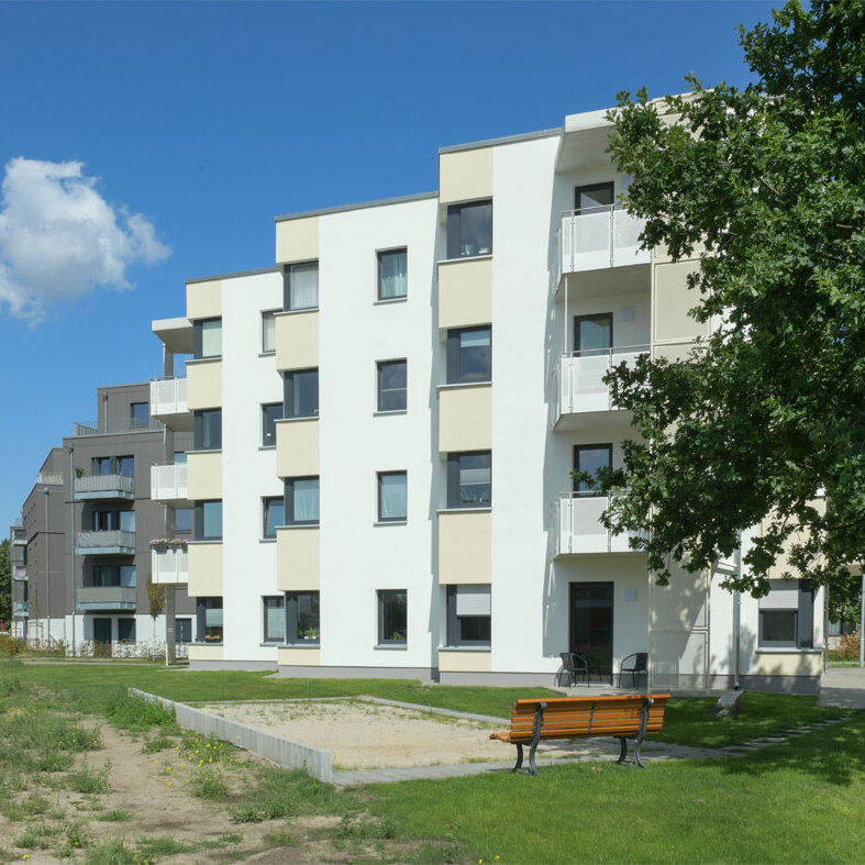 Außenansicht degewo-Neubau Wilhelm-Hoff-Straße 24 und 26 mit Grünfläche, Sandkasten, Sitzbank und Baum rechts im Vordergrund.