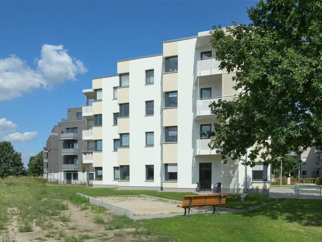 Außenansicht degewo-Neubau Wilhelm-Hoff-Straße 24 und 26 mit Grünfläche, Sandkasten, Sitzbank und Baum rechts im Vordergrund.