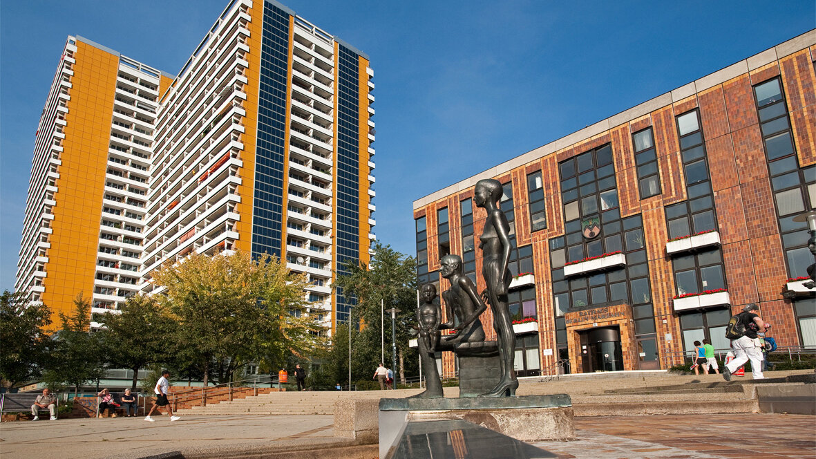 Öffentlicher Platz mit einer Figurengruppe aus Bronze im Vordergrund, dahinter ein markantes Hochhaus mit gelben Fassadenelementen und ein weiteres Gebäude mit Backsteinfassade.