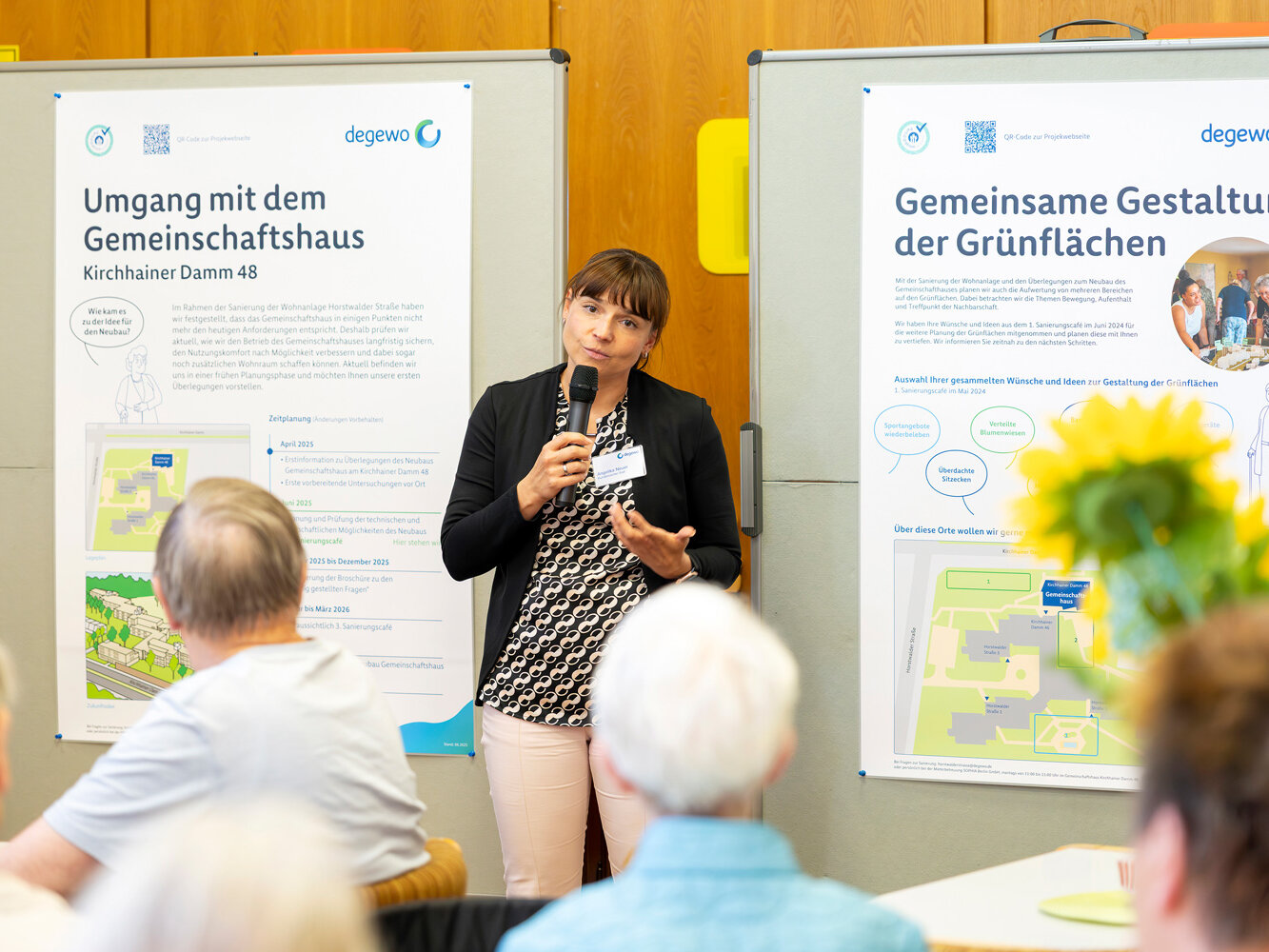 Eine Projektmitarbeiterin spricht mit Mikrofon vor einer Gruppe von Teilnehmenden. Hinter ihr stehen Infoplakate zur Gestaltung der Grünflächen und zum Gemeinschaftshaus.