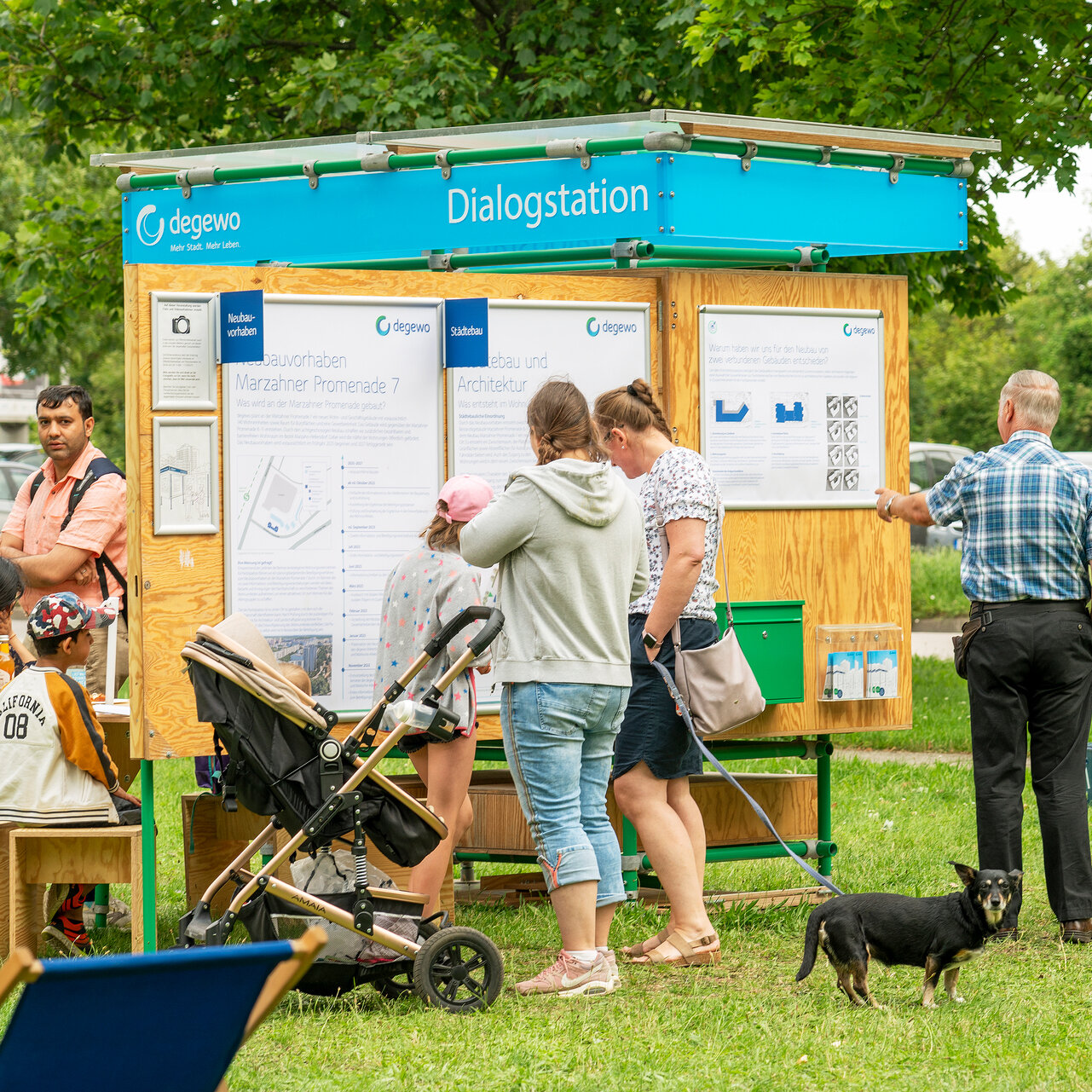 Die degewo-Dialogstation steht auf einer grünen Wiese. An den aufgeklappten Seiten hängen Plakate. Davor stehen Personen und schauen sich die Plakate an. An der hinteren Seite der Dialogstation sitzen weitere Personen an einem kleinen Tisch.
