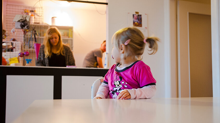 Kleinkind sitzt am Tisch und schaut nach hinten zur Mutter.