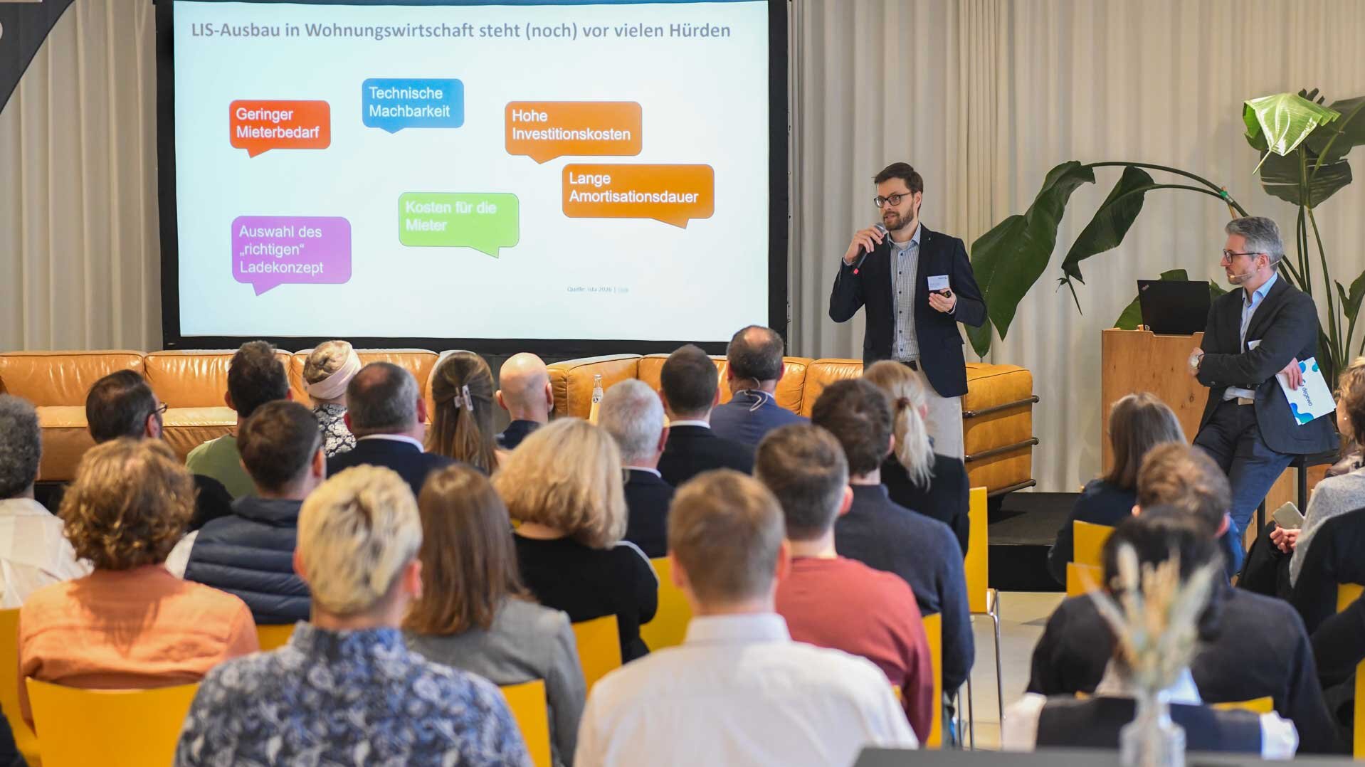 Das Publikum schaut Richtung Bühne. Dort steht ein Mann und hält einen Vortrag. Im Hintergrund auf einer Leinwand ist eine Präsentation zu sehen.