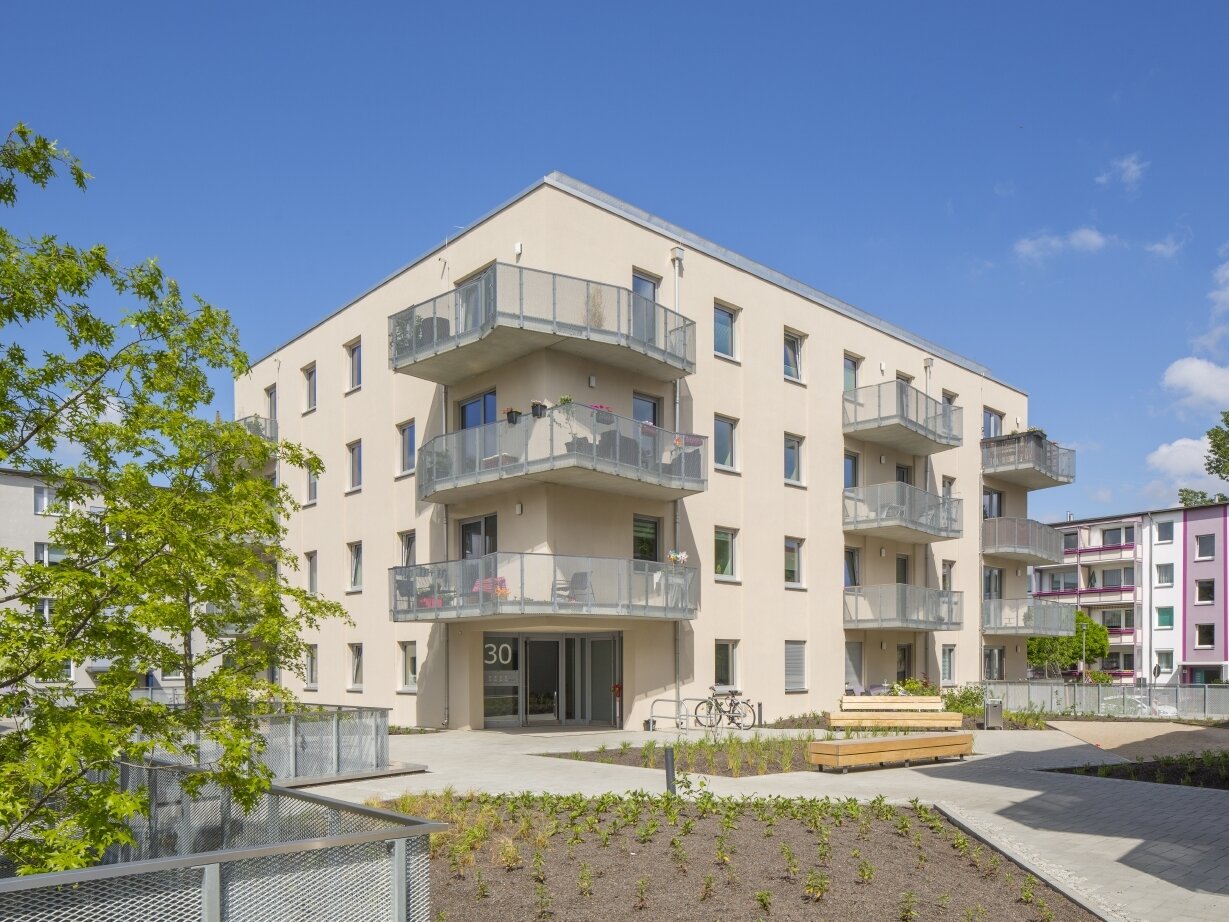 Außenansicht degewo-Neubau Karl-Pokern-Straße 30 mit Bäumen und Zäunen links sowie Beeten, Wegen und Sitzbänken im Vordergrund.