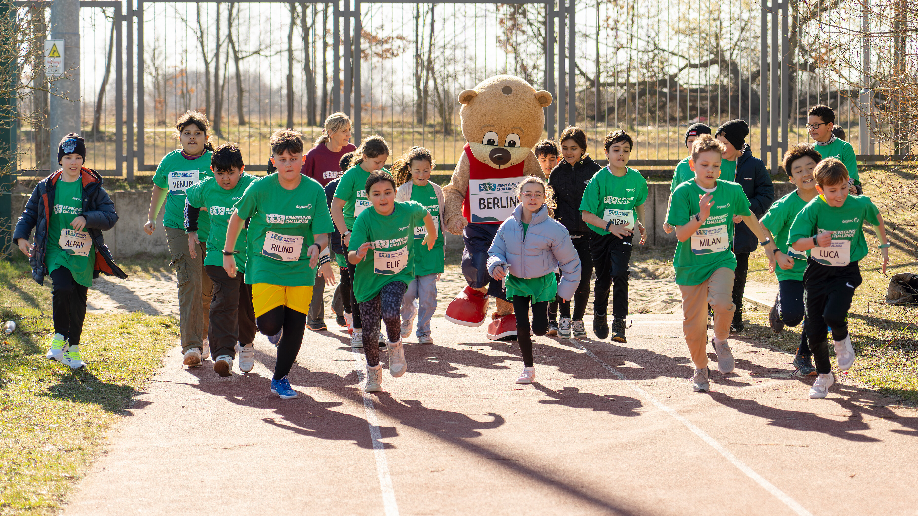 Kinder laufen bei einem Rennen auf einer Bahn, begleitet von einem Bären-Maskottchen.