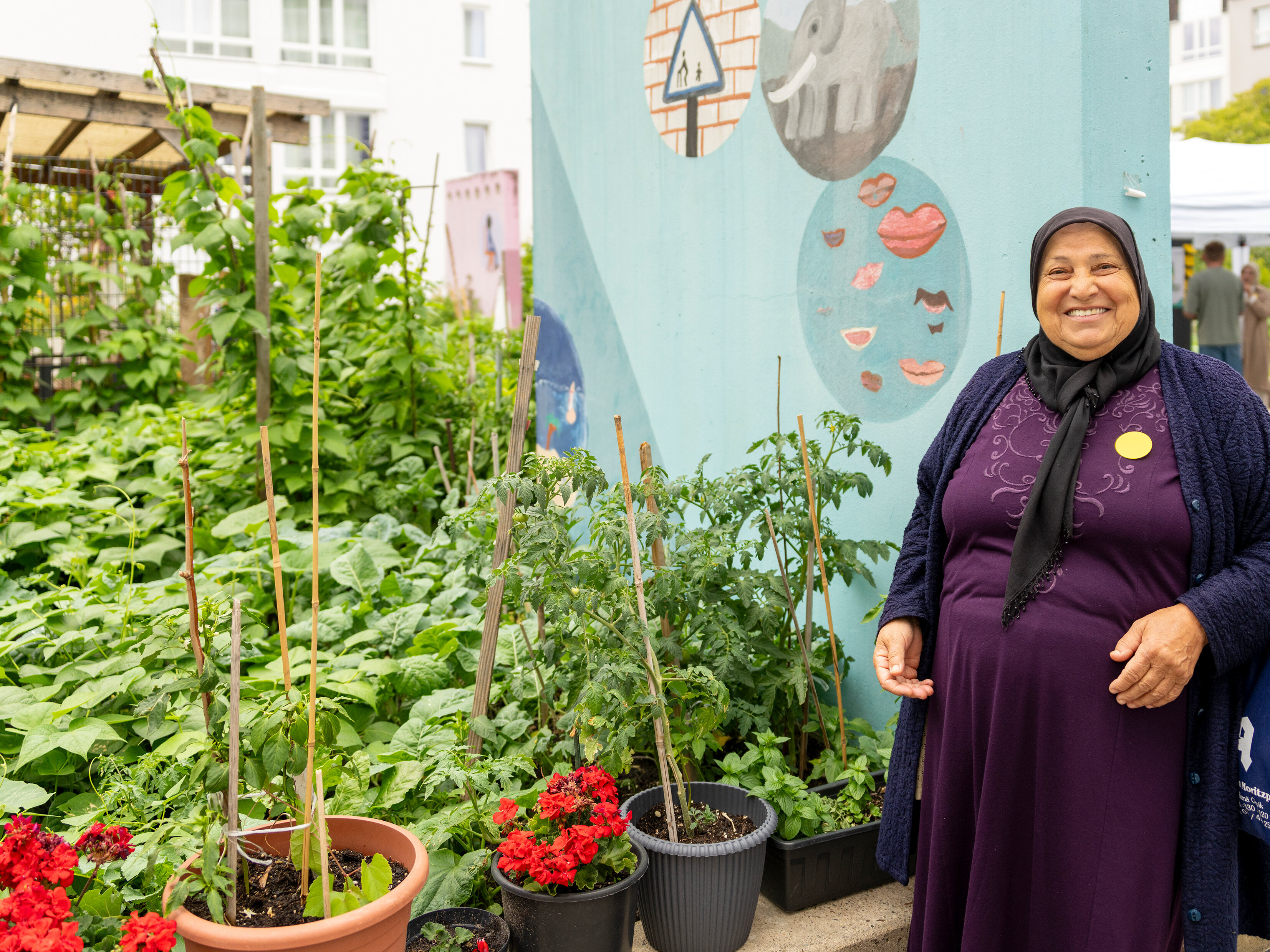 Teilnehmerin lächelt vor bepflanzten Hochbeeten im Innenhof. Hinter ihr farbig gestaltete Wandelemente und ein gemeinschaftlich genutzter Gartenbereich.