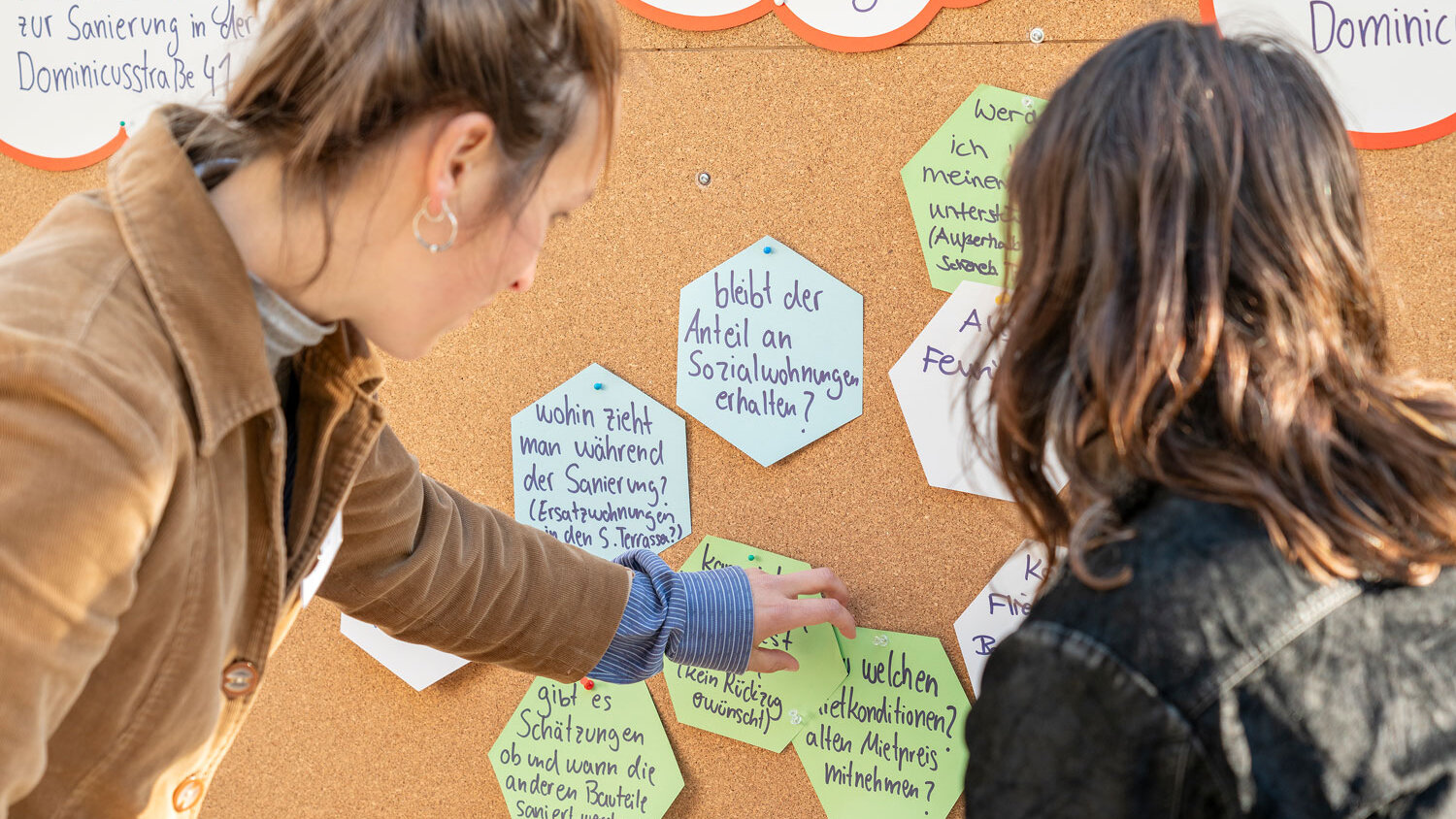 Zwei Frauen heften handgeschriebene Fragen zur Sanierung an eine große Pinnwand. Die Zettel haben unterschiedliche Formen und Farben.