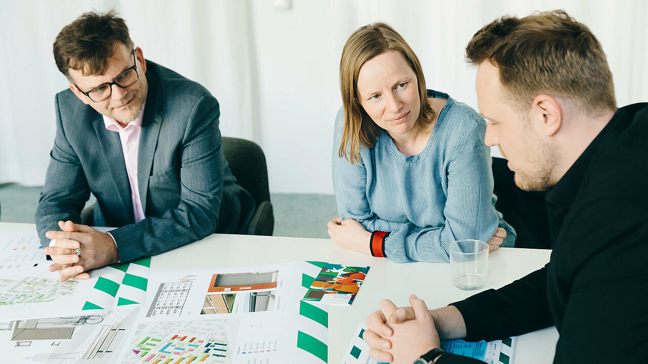 Drei Kollegen sitzen am Tisch und diskutieren mit Plänen vor sich liegend.