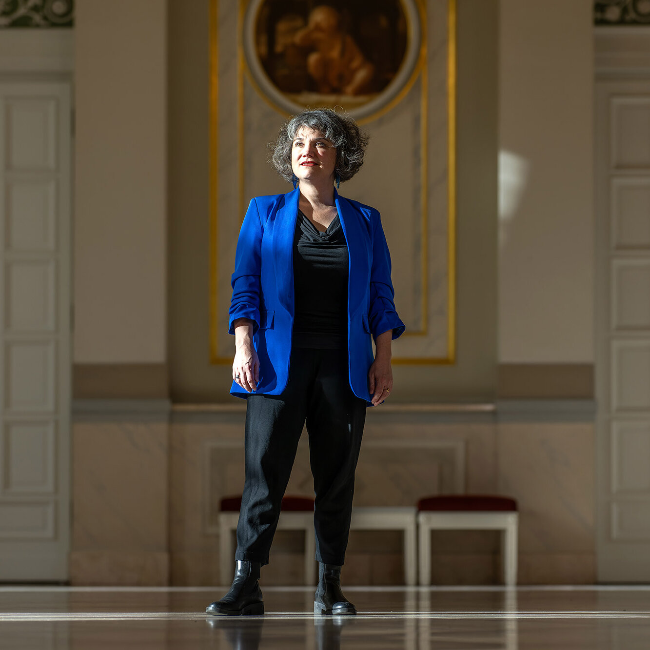 Eine Frau steht in einem großen historischen Saal und trägt ein blaues Sakko. Sie blickt leicht nach oben und wird vom Sonnenlicht angestrahlt.