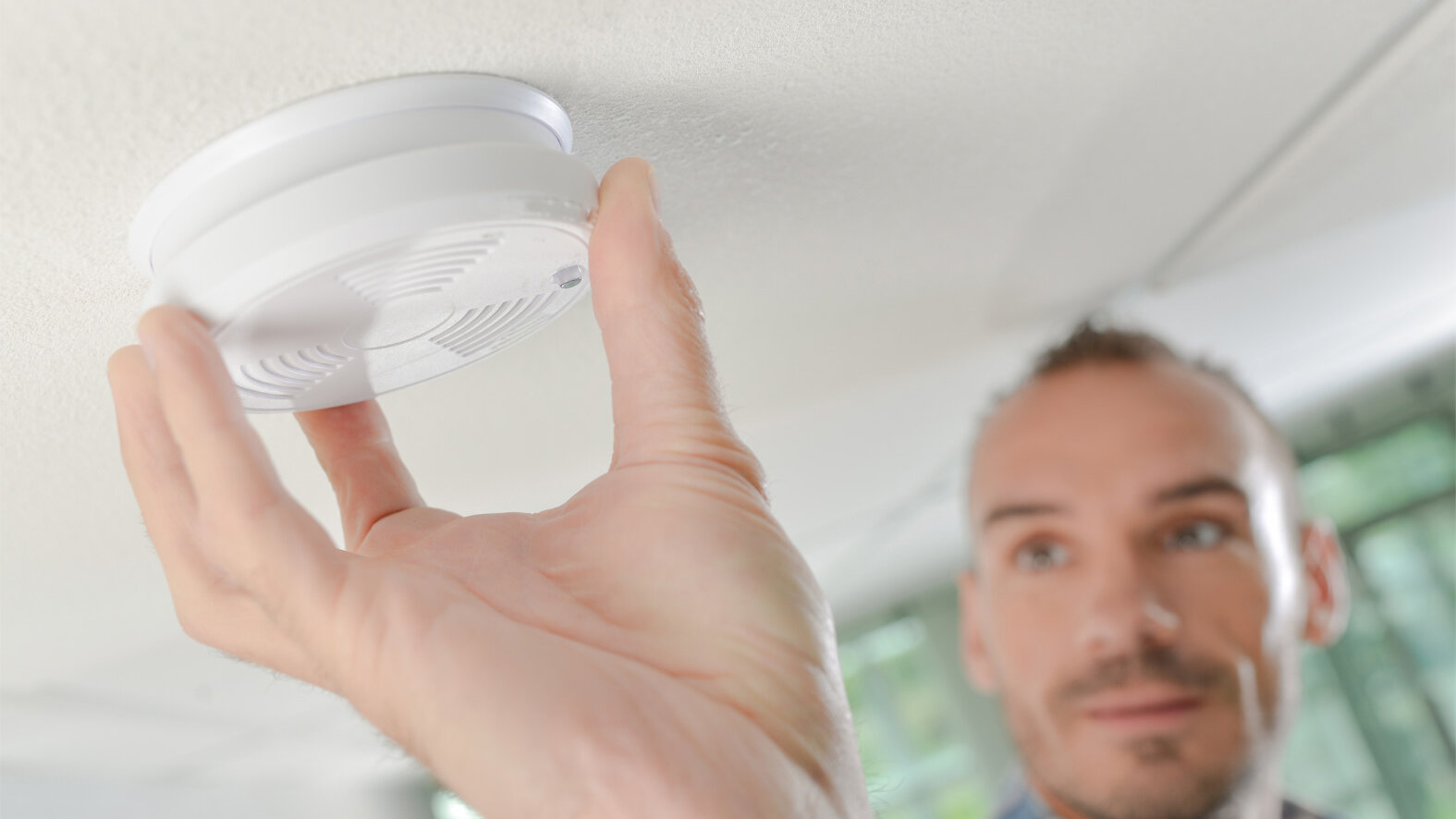 Person montiert oder überprüft einen Rauchwarnmelder an der Zimmerdecke. Der Rauchmelder wird mit der Hand an der Halterung befestigt.