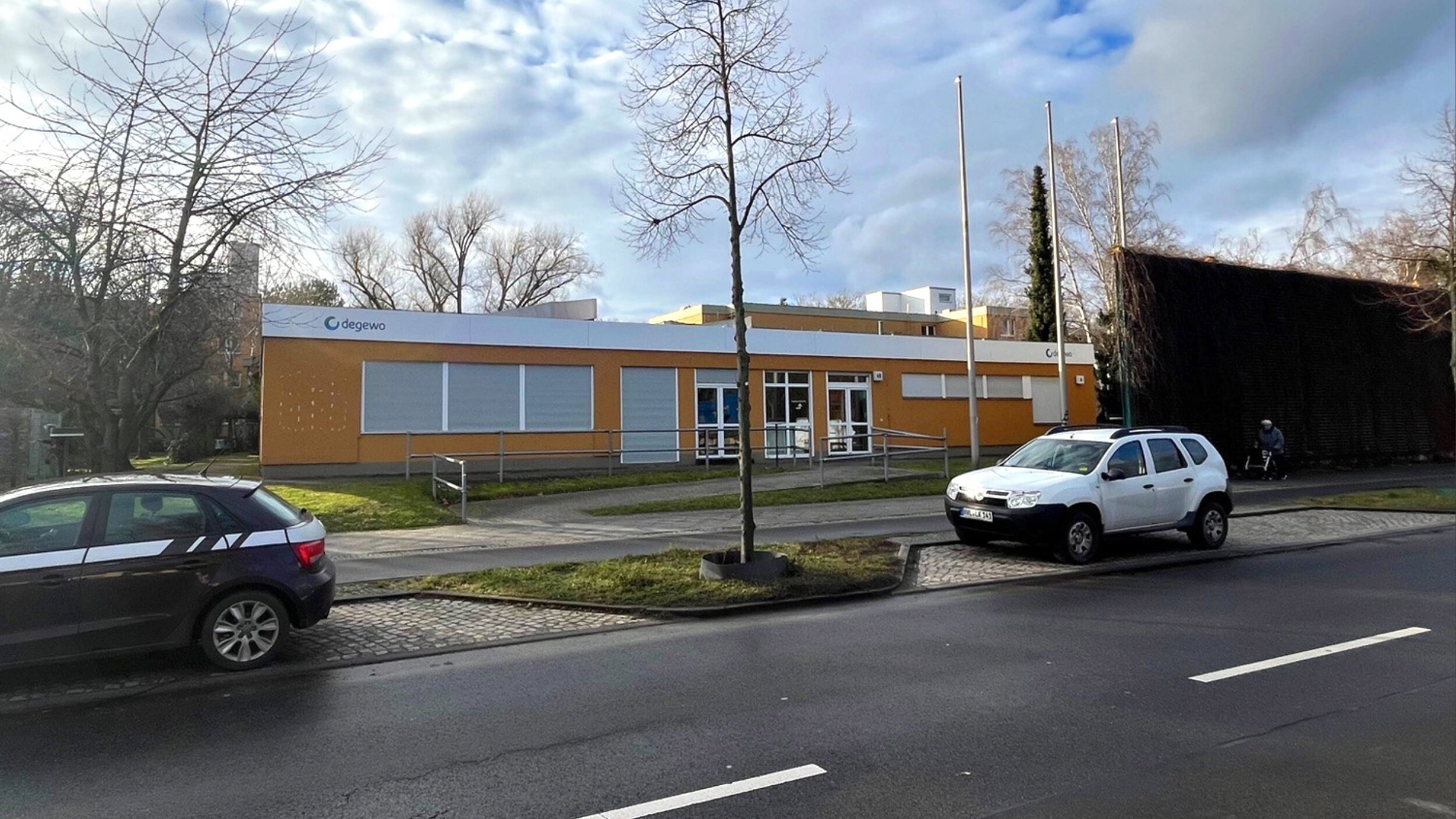 Außenansicht des Gemeinschaftshauses Horstwalder Straße: eingeschossiges, orangefarbenes Gebäude mit degewo-Logo, barrierefreiem Eingang und Rampe, davor Straße mit parkenden Autos und winterlichen Bäumen.
