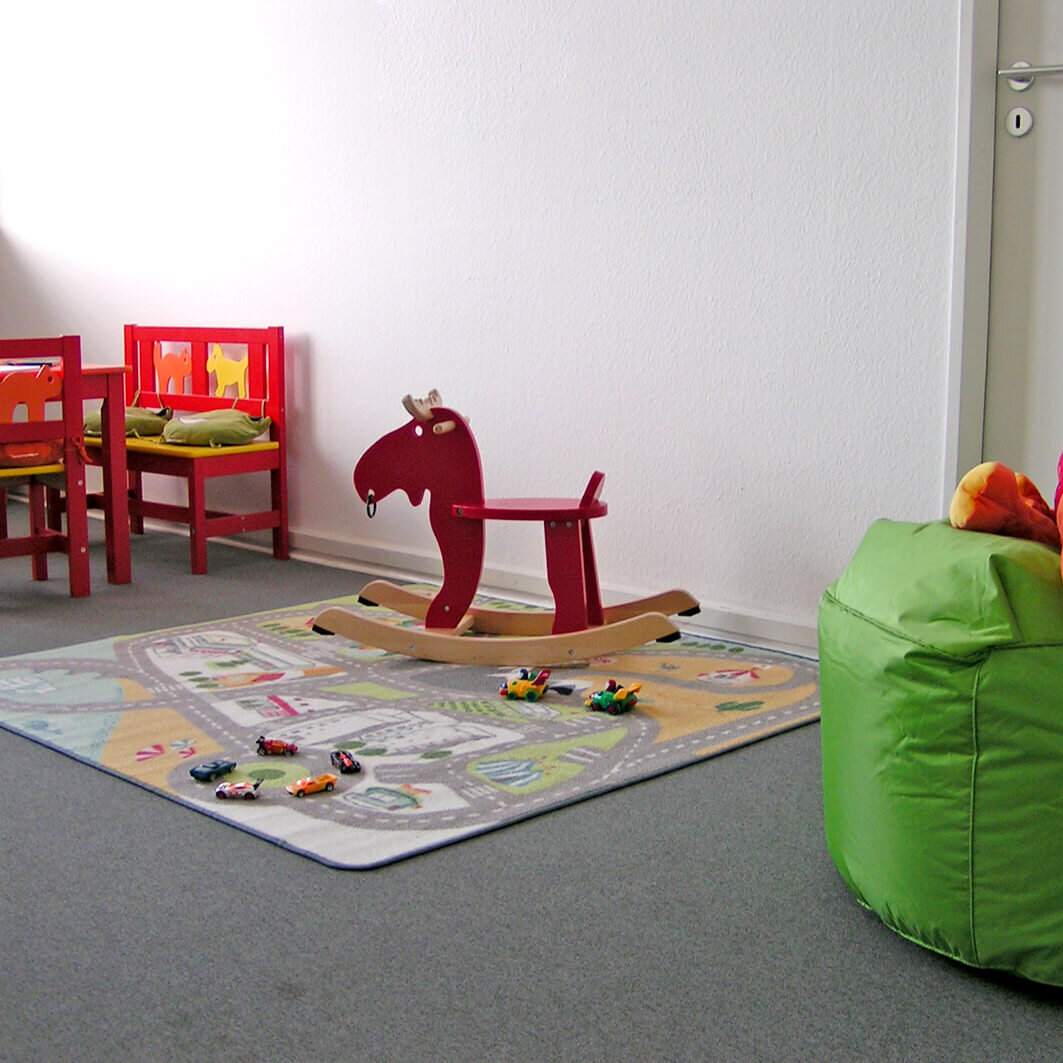 Heller Spielraum mit kleinem Tisch und Stühlen, Schaukelpferd, Spielteppich mit Autos und grünem Sitzsack