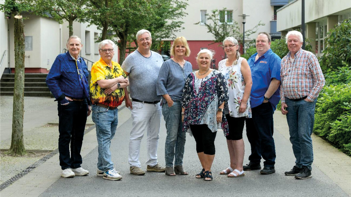 Gruppenaufnahme des Mieterbeirates. Acht Personen stehen im Freien auf einem Hof. Im Hintergrund ist ein Wohnhaus zu sehen. 